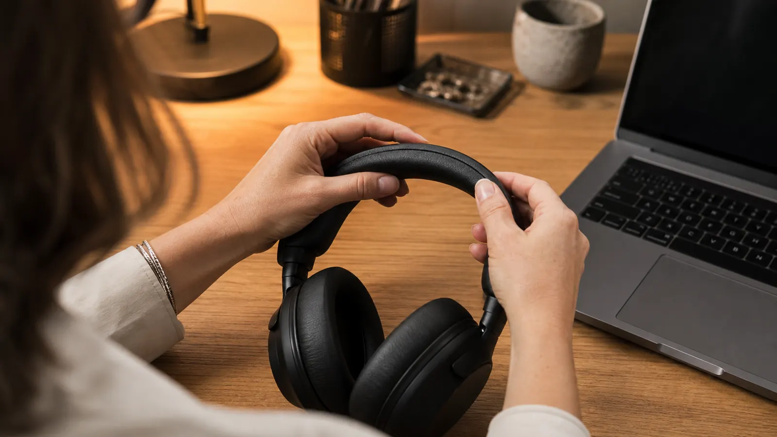 Close-up of hands adjusting the headband of black over-ear noise cancelling headphones at a desk