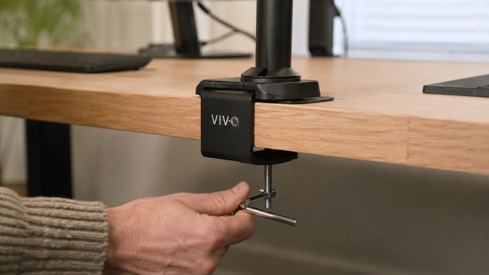 A hand tightening the desk clamp of a monitor arm onto the back edge of a wood desk