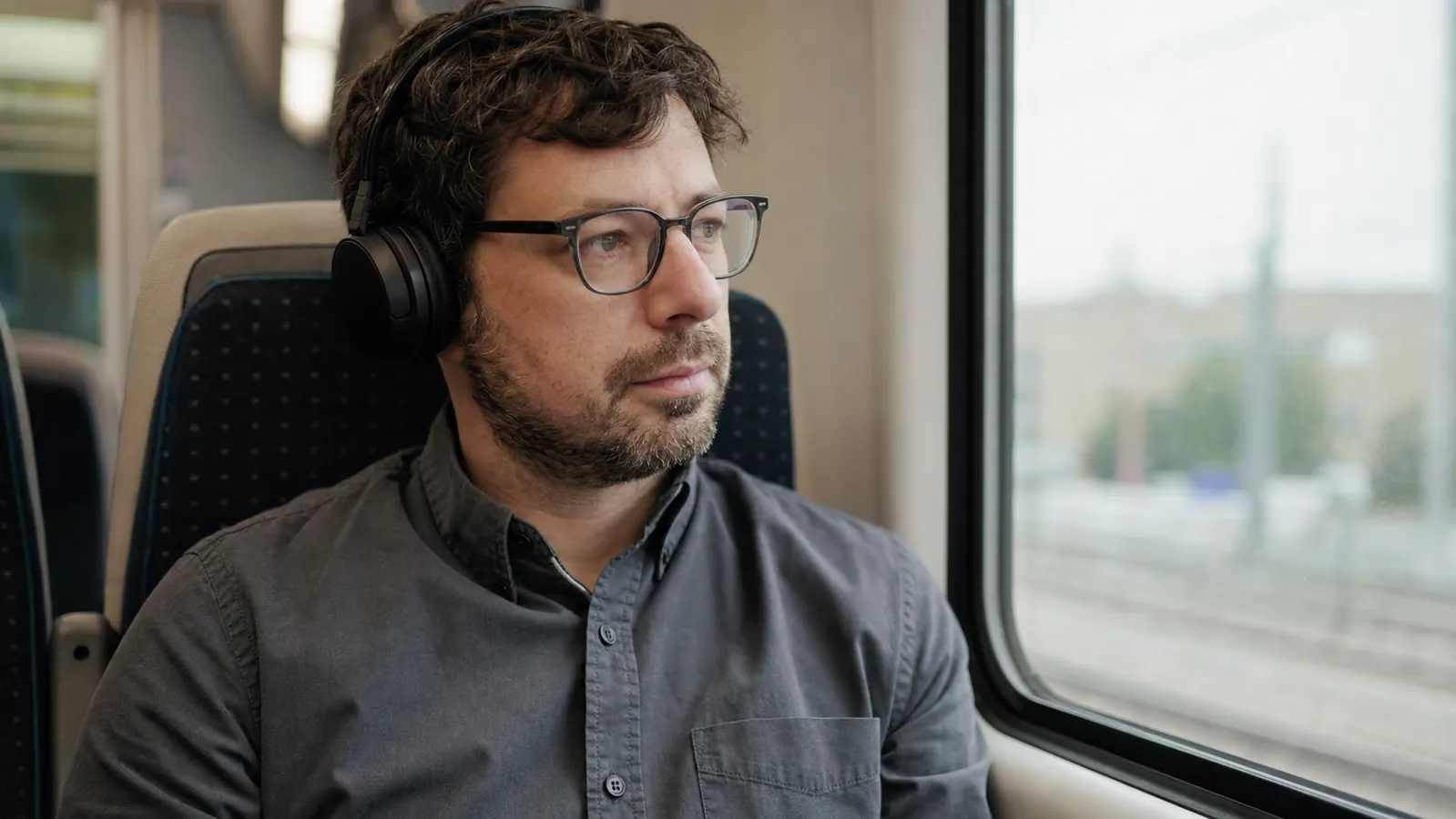 Commuter wearing glasses and over-ear headphones seated on a train looking out the window