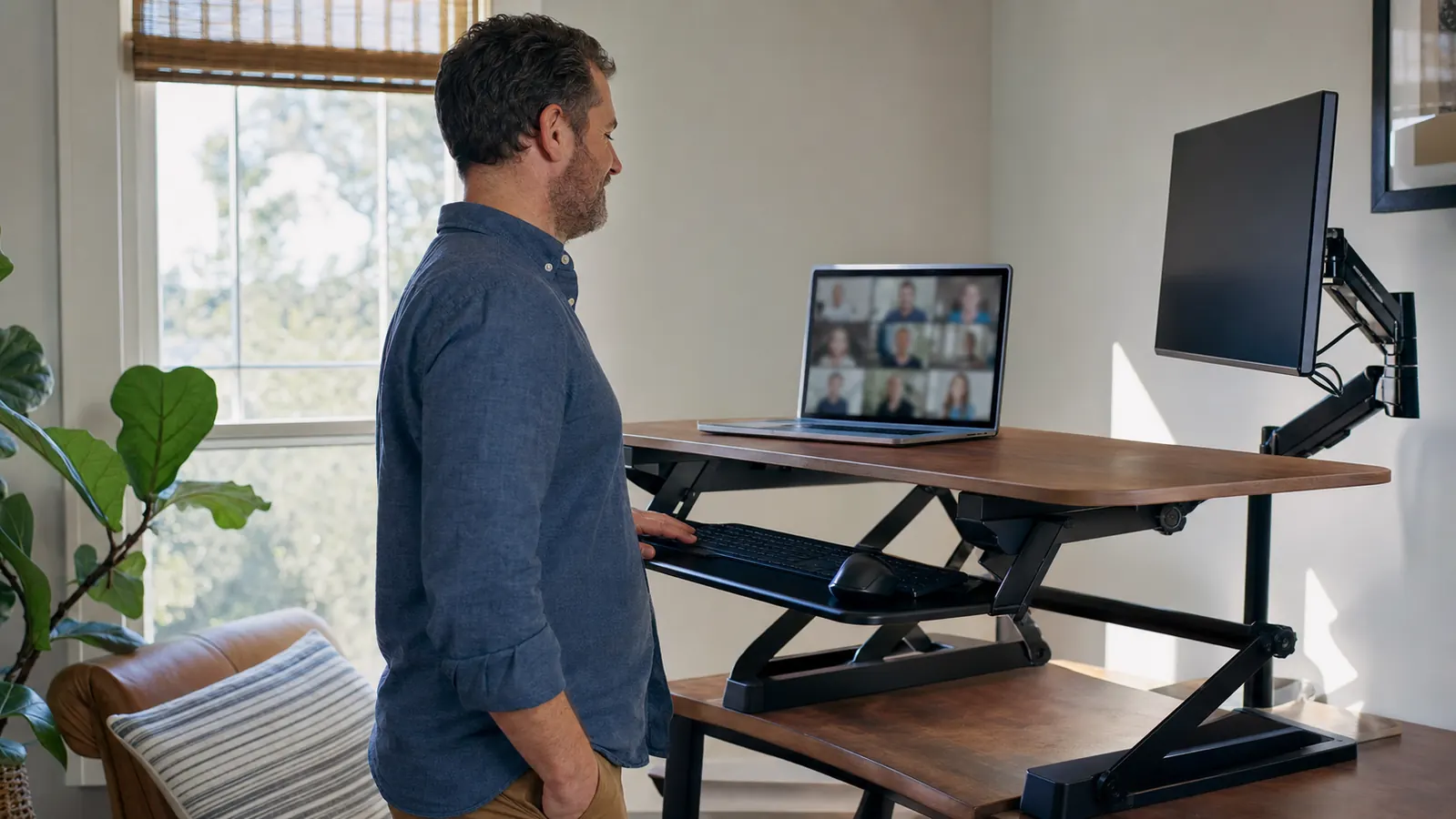 Home office worker standing at a raised desk converter mid video call, monitor showing a generic call grid