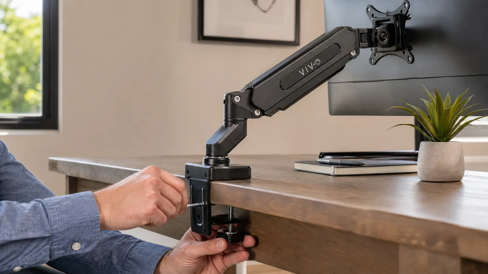Close-up of a hand tightening the clamp of a monitor arm to the edge of a wooden desk