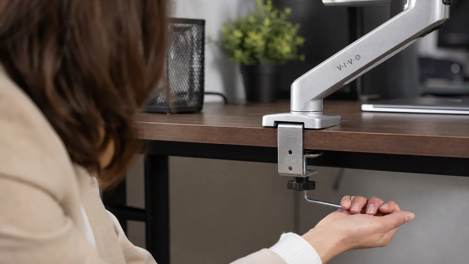 Hand tightening the C-clamp base of a monitor arm to the back edge of a wooden desk