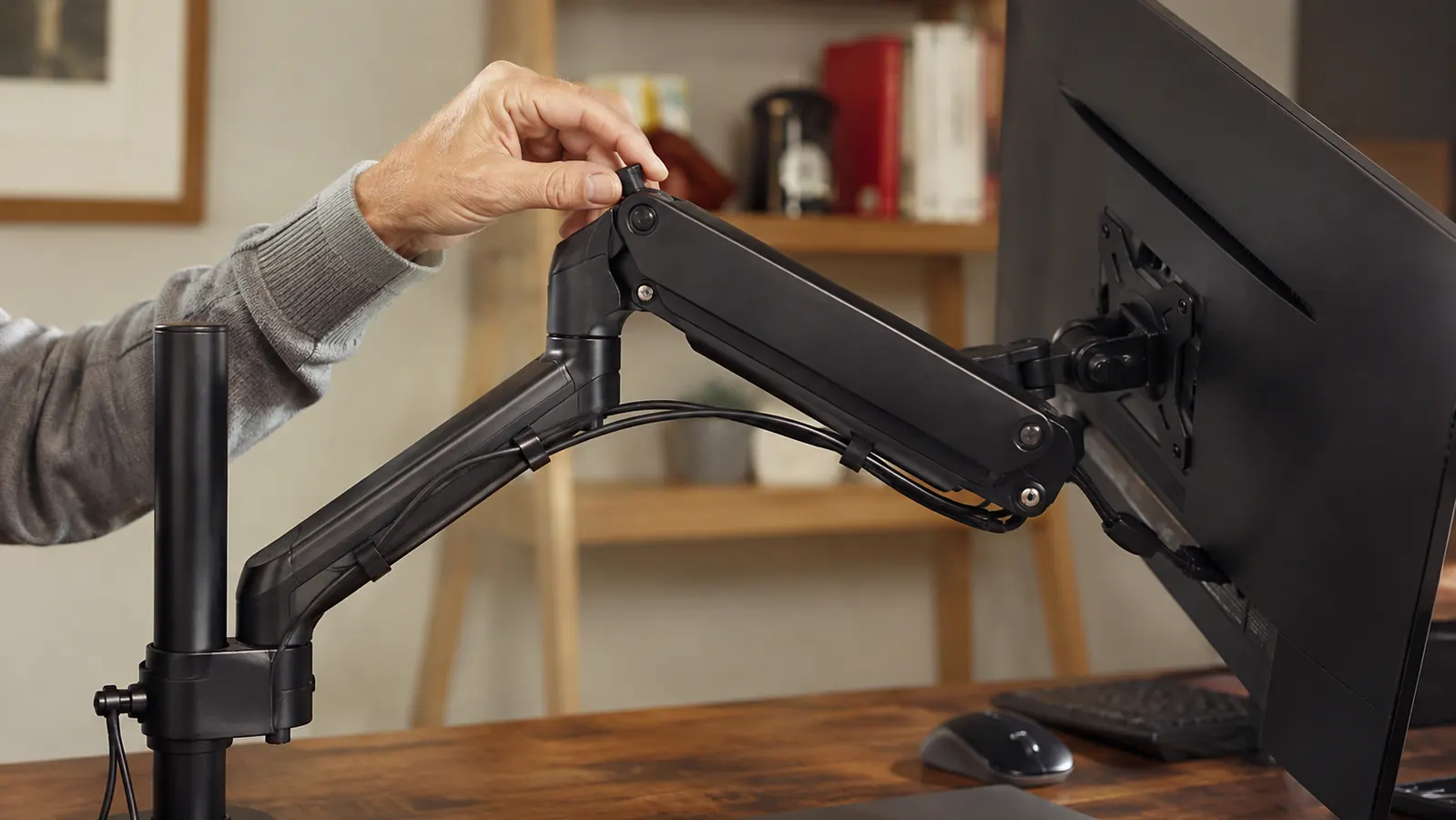 Close-up of a hand adjusting the tension knob on a black articulating monitor arm clamped to the back edge of a desk, monitor swung out to the side